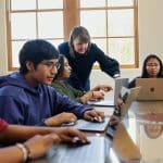 Teacher guiding students using laptops in a classroom setting.