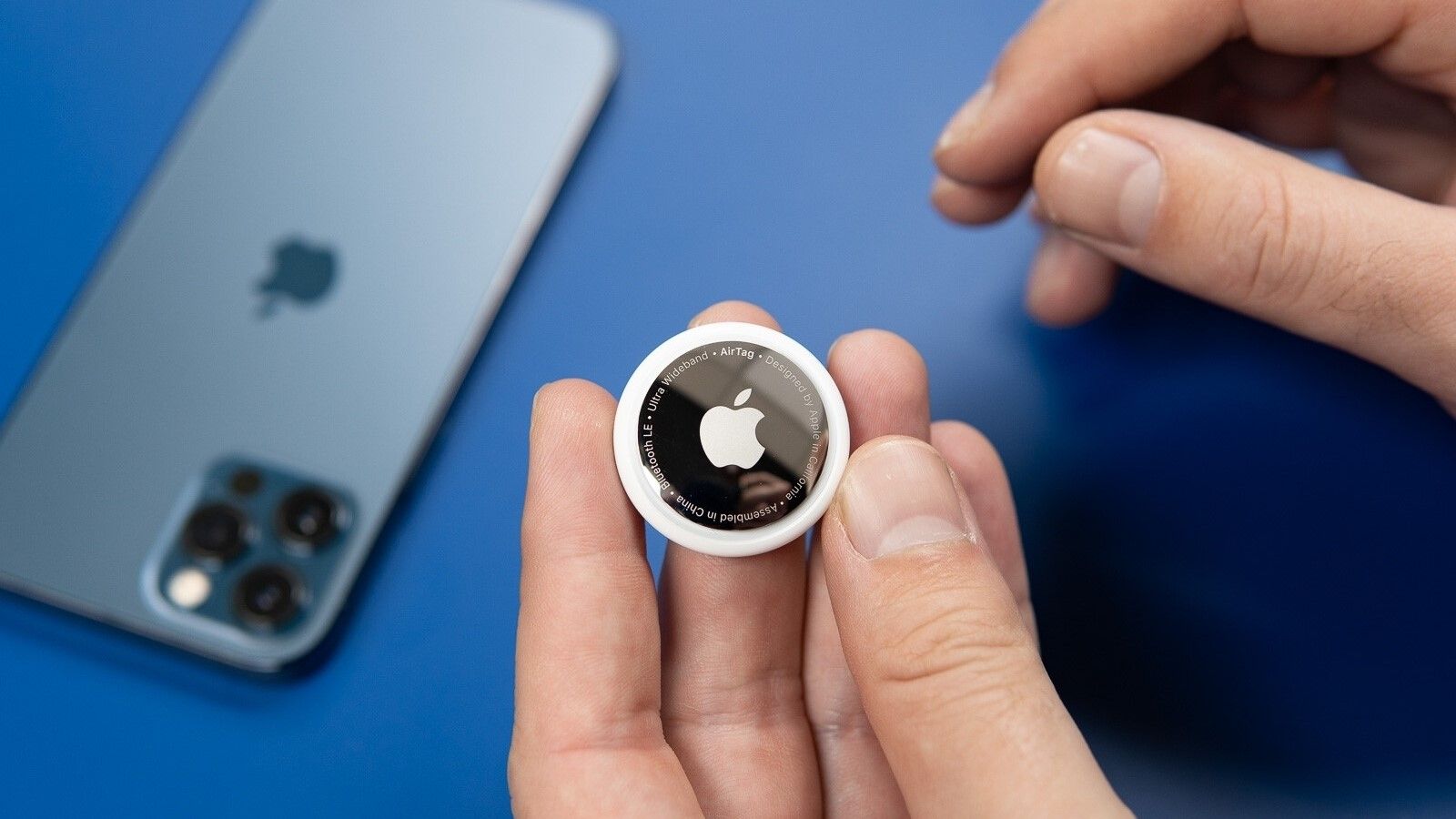 Person holding an Apple AirTag with an iPhone in the background.